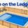 A fenced pool has a shallow ledge, closed gate, and water shoes outside.